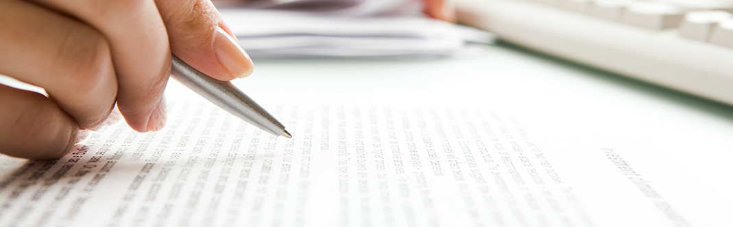 A person's hand holding a pen over a printed document, with focus on the text and a blurred background featuring a keyboard and papers