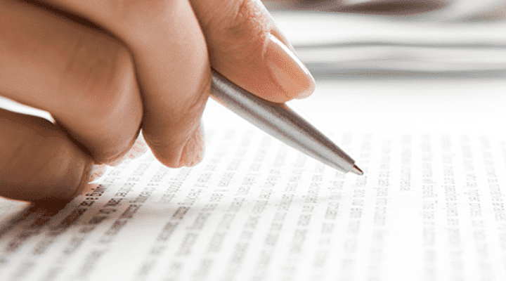 A person's hand holding a pen over a printed document, with focus on the text and a blurred background featuring a keyboard and papers